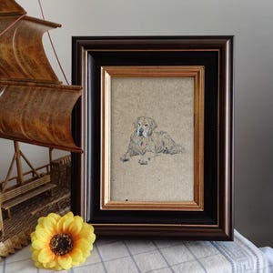 May include: A framed drawing of a golden retriever dog lying down. The dog is looking directly at the viewer. The drawing is on a beige background and is framed in a black and gold frame.