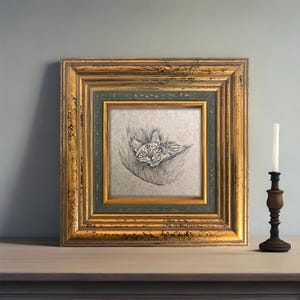 May include: A framed pencil sketch of a cat resting in a hammock. The artwork is in a gold and green ornate frame. A wooden candle holder with a white candle is to the right of the frame.