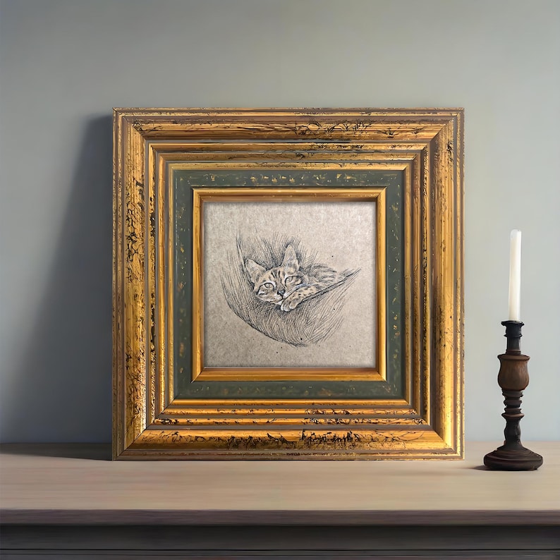 May include: A framed pencil sketch of a cat resting in a hammock. The artwork is in a gold and green ornate frame. A wooden candle holder with a white candle is to the right of the frame.