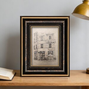 May include: A rectangular picture frame with a black and gold ornate design. The frame holds a pencil sketch of a building with a cafe. The frame is sitting on a wooden surface.
