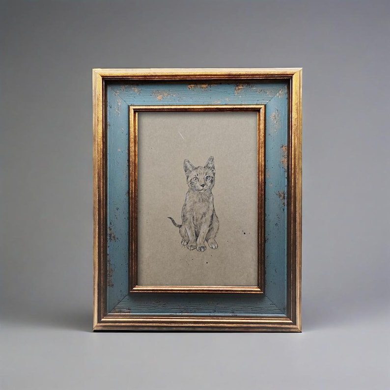 May include: A framed pencil sketch of a seated cat. The frame has a distressed blue finish with gold trim. The artwork is on a tan background, creating a vintage aesthetic. The cat is drawn in detail, with visible fur and facial features.