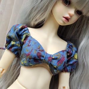 May include: A blue and burgundy floral print doll top with puffed sleeves. The top has a bow detail in the front.