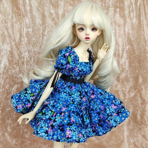 May include: A doll wearing a blue and white floral dress with a black ribbon and lace trim. The doll has long blonde hair and is wearing a white wig.