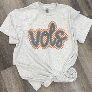 May include: A grey t-shirt with the word "VOLS" in black outlined in white and orange checkered pattern.