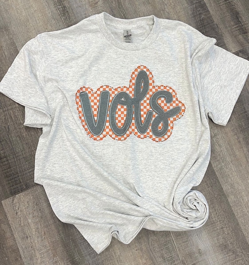 May include: A grey t-shirt with the word "VOLS" in black outlined in white and orange checkered pattern.