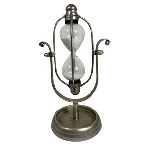 May include: A vintage hourglass with a clear glass bulb filled with white sand, encased in a silver-toned metal frame. The hourglass sits on a round base and has decorative curved accents. A classic timekeeping device.