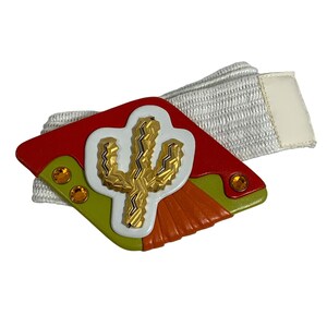 May include: A colorful belt with a decorative buckle featuring a gold cactus design. The buckle has red, green, orange, and white accents with two orange gemstones. The belt is white and has a cream-colored tab.