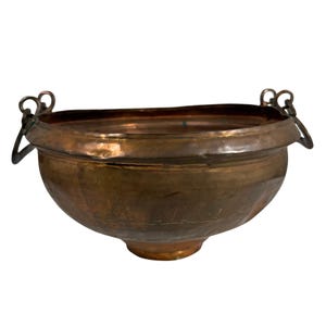 May include: A round, hammered copper bowl featuring a wide, flared rim and two metal ring handles attached to the sides. The bowl has a deep, warm metallic finish with visible signs of wear and a small, circular base at the bottom center.