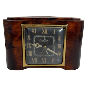 May include: A vintage Sessions clock with a dark wood finish and a rectangular face. The clock face is black with gold Roman numerals and hands. The clock has a gold-colored frame and the Sessions logo.
