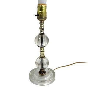 May include: A clear glass table lamp with a round base and two spherical glass accents. The lamp has a brass and silver metal stem and a standard light bulb socket. A brown electrical cord extends from the base.