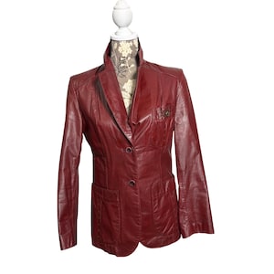 May include: A burgundy leather blazer displayed on a mannequin. The blazer features a notched lapel, two front pockets, and a two-button closure. The jacket has long sleeves and a tailored fit, showcasing a classic style.