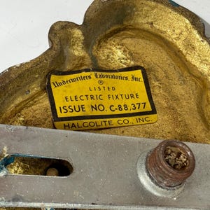 May include: Close-up of a vintage electric fixture component. The fixture has a gold-colored, ornate design with teal accents. A yellow label reads "Underwriters Laboratories, Inc." and "Electric Fixture." A metal bracket and a rusty screw are also visible.