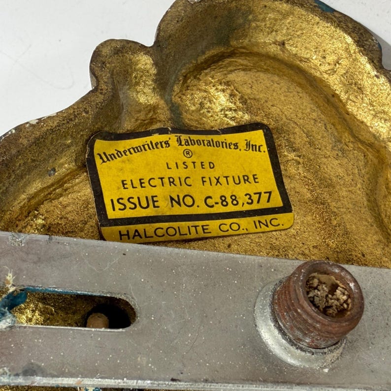 May include: Close-up of a vintage electric fixture component. The fixture has a gold-colored, ornate design with teal accents. A yellow label reads "Underwriters Laboratories, Inc." and "Electric Fixture." A metal bracket and a rusty screw are also visible.
