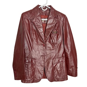 May include: A dark burgundy leather blazer featuring a notched lapel, two front button closures, and three smaller buttons on each cuff. The jacket includes two lower patch pockets and one chest pocket with a gold-toned metal hardware detail.