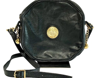 Puede incluir: Bolso bandolera de cuero negro con un emblema circular dorado en la parte delantera. El bolso tiene una correa larga y ajustable con una hebilla dorada y detalles de cadena dorada con pequeños dijes de bola. Forma octogonal.