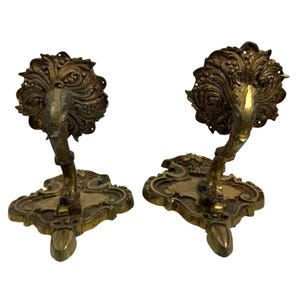 May include: Two antique brass wall sconces. Each sconce features an ornate, floral design with a curved arm and a decorative base. The sconces are made of a gold-toned metal and are likely vintage.