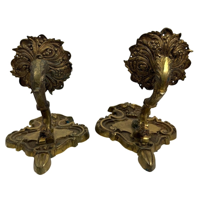 May include: Two antique brass wall sconces. Each sconce features an ornate, floral design with a curved arm and a decorative base. The sconces are made of a gold-toned metal and are likely vintage.