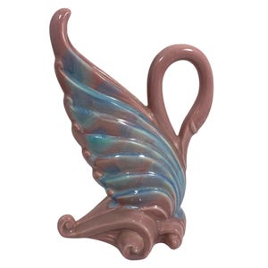 May include: A ceramic decorative vase in a wing-like shape. The vase features a mauve base and handle, with a wing design in shades of blue and teal. The handle is a loop shape. The vase has a glossy finish.