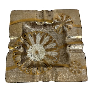 May include: Square glass ashtray with a gold glitter background and gold floral designs. The ashtray has a textured surface and a wavy edge. The floral designs are in the center and on the sides.
