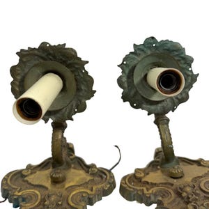 May include: Two antique brass wall sconces. Each sconce features a decorative base with ornate detailing, a curved arm, and a floral-shaped backplate. The sconces have a vintage aesthetic, with visible wear and a warm, aged patina.