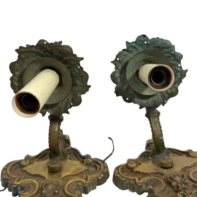 May include: Two antique brass wall sconces. Each sconce features a decorative base with ornate detailing, a curved arm, and a floral-shaped backplate. The sconces have a vintage aesthetic, with visible wear and a warm, aged patina.