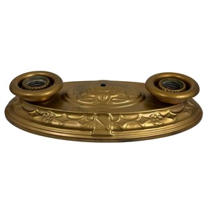 May include: An oval-shaped, metallic gold-toned light fixture base featuring two circular bulb sockets on the top surface. The base is decorated with embossed floral and geometric patterns along the edges and a central raised oval design.