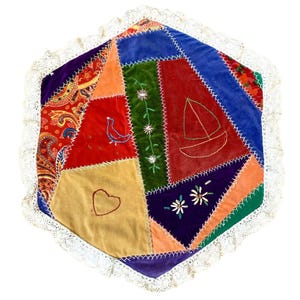 May include: A colorful, patchwork cushion with a white lace trim. The cushion features various velvet patches in red, blue, green, orange, and gold, with embroidered details including a bird, sailboat, and flowers. The cushion is approximately 16 inches in diameter.