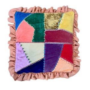 May include: A square patchwork pillow with a ruffled edge. The pillow features a variety of velvet squares in shades of blue, green, gold, yellow, red, purple, and peach. The squares are stitched together with white thread and accented with small beads.