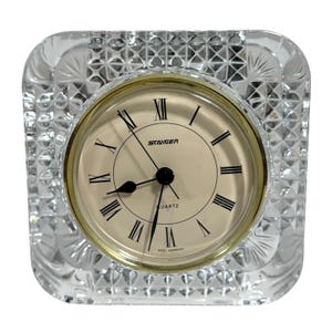 May include: A clear, square-shaped crystal clock with a gold-tone bezel and a cream-colored face. The clock face features black Roman numerals and the brand name "STAIGER". The clock is marked "QUARTZ" and "WEST GERMANY".