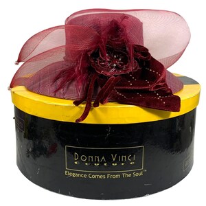 May include: A burgundy hat with a wide brim and decorative accents, including a velvet flower and feathers. The hat is displayed on a black and yellow round box. The box has the text "Donna Vinci Couture" and "Elegance Comes From The Soul".