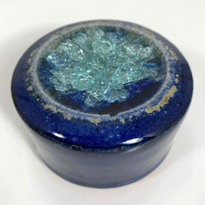 May include: A round, ceramic container with a deep blue exterior and a light blue interior filled with crushed glass. The rim has a textured, off-white and gold trim. The container is a decorative item.