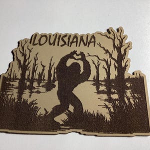 May include: A wooden cutout of the state of Louisiana featuring a silhouette of a Bigfoot-like creature in a swamp scene. The word "LOUISIANA" is at the top. The cutout is brown with a lighter tan background.