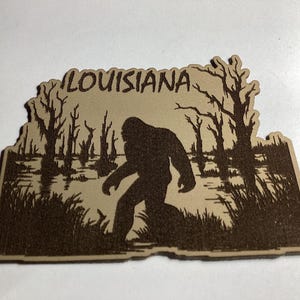 May include: A wooden cutout featuring a silhouette of Bigfoot in a swamp scene. The word "LOUISIANA" is etched at the top. The design is in shades of brown and beige, depicting trees, water, and tall grass.