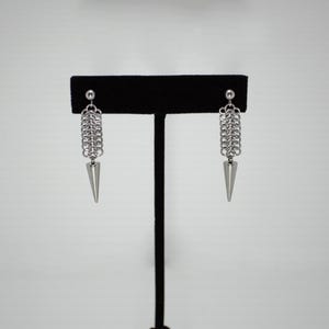 Puede incluir: Un par de pendientes de plata con un diseño de cadena y una punta puntiaguda. Los pendientes se muestran en un soporte negro.