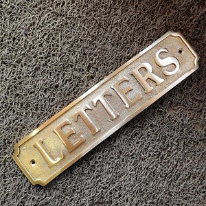 May include: A brass sign with rounded edges and a black dot in the top right corner. The sign reads "LETTERS" in raised white letters.