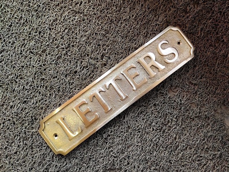 May include: A brass sign with rounded edges and a black dot in the top right corner. The sign reads "LETTERS" in raised white letters.