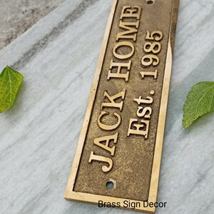 May include: A brass sign with the text "JACK HOME Est. 1985" in a vintage style font. The sign has a distressed finish and a hole in the top for hanging.