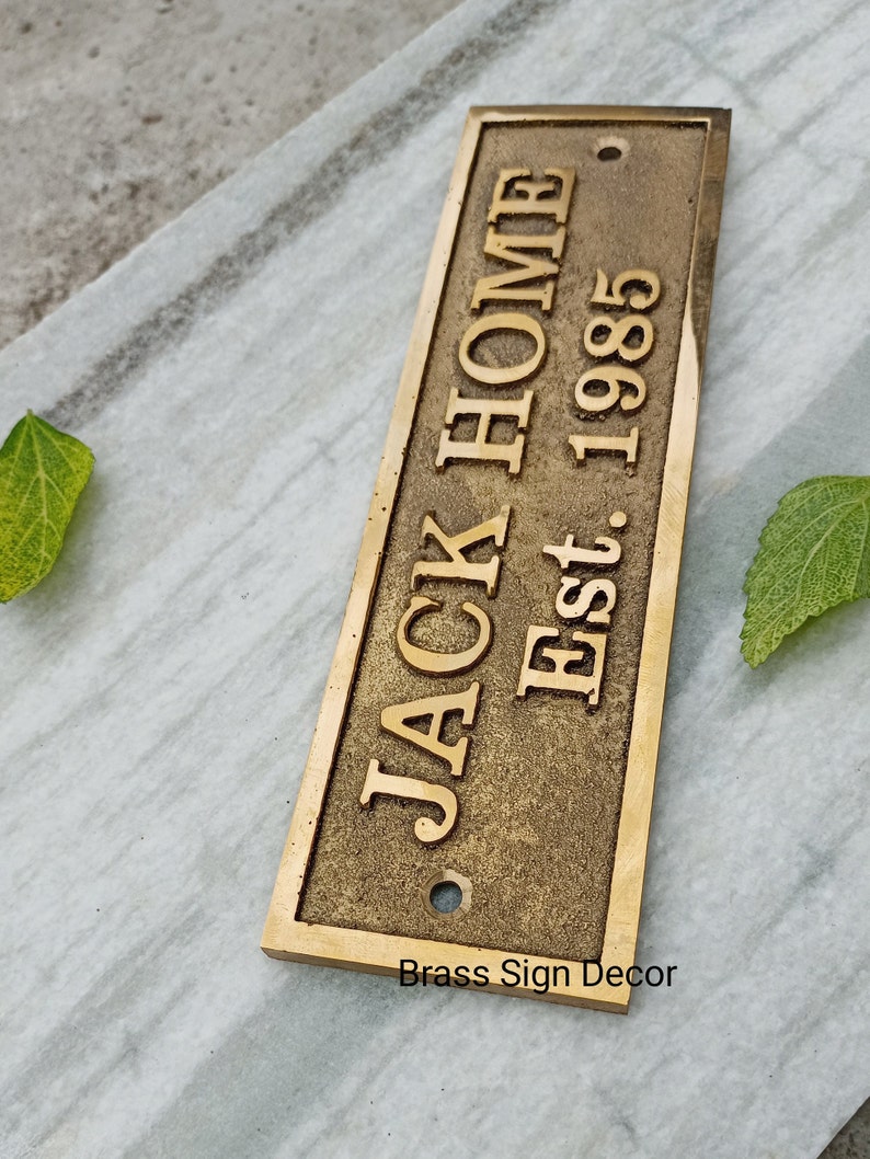 May include: A brass sign with the text "JACK HOME Est. 1985" in a vintage style font. The sign has a distressed finish and a hole in the top for hanging.