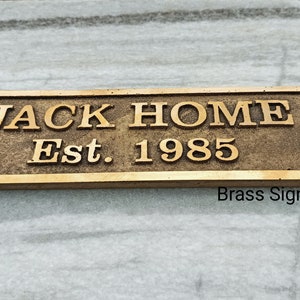 May include: A brass sign with the text "JACK HOME Est. 1985" on a white surface.