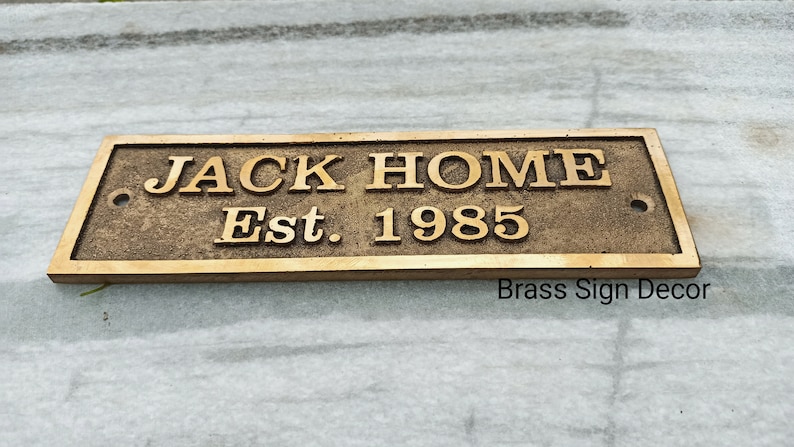 May include: A brass sign with the text "JACK HOME Est. 1985" on a white surface.