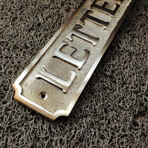 May include: A brass sign with the word "LETTER" engraved on it. The sign has a rounded edge and a hole in the centre.
