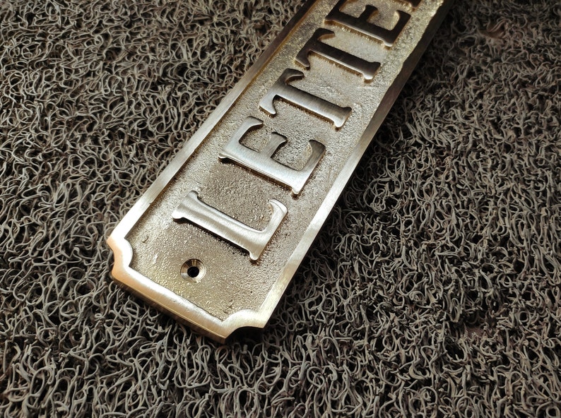 May include: A brass sign with the word "LETTER" engraved on it. The sign has a rounded edge and a hole in the centre.