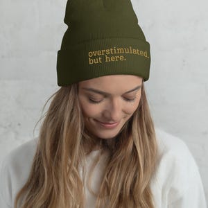 Op de afbeelding: Een olijfgroene muts met omslag en de geborduurde tekst "overstimulated. but here." in een lichtbeige kleur. De gebreide muts heeft een geribbelde textuur en een omgeslagen rand, wat zorgt voor een eenvoudig en functioneel ontwerp voor hoofddeksels.