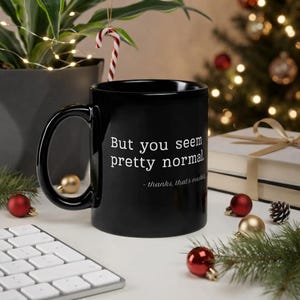 Könnte beinhalten: Ein schwarzer Keramikbecher mit weißem Text, der lautet: "But you seem pretty normal. - thanks, that's masking." Der Becher hat einen geschwungenen Henkel und eine glatte Oberfläche und dient als funktionaler Behälter für heiße oder kalte Getränke.