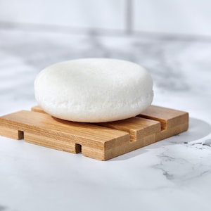 Può includere: Una saponetta bianca e rotonda poggia su un portasapone in legno naturale. Il portasapone ha una forma rettangolare con fessure di drenaggio. Lo sfondo è una superficie marmorizzata.