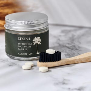 May include: A jar of "DESESH" 60 whitening toothpaste tablets with a natural mint flavor. A wooden toothbrush with black bristles and several white tablets are next to the jar. The jar has a silver lid and a dark green label with a palm tree illustration.