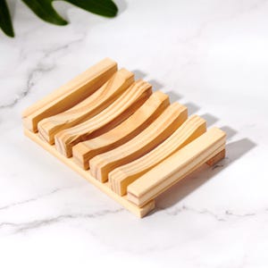 Op de afbeelding: Een lichtgekleurde houten zeepbakje met een rechthoekige basis en gebogen latten. Het bakje is gemaakt van natuurlijk hout en heeft een eenvoudig, minimalistisch ontwerp. Het zeepbakje is ongeveer 13 cm lang en 8 cm breed.