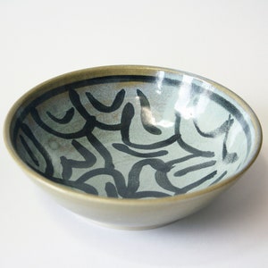 Könnte beinhalten: Eine Keramikschüssel mit hellgrüner Glasur und einem schwarzen abstrakten Design. Die Schüssel ist rund und hat einen leichten Rand.
