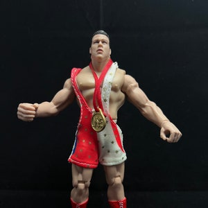Puede incluir: Una figura de acción de lucha libre roja, blanca y azul con una medalla de oro alrededor del cuello. La figura lleva un pantalón de lucha libre rojo y blanco con estrellas y una bata de lucha libre roja y blanca. La figura está de pie sobre un fondo negro.