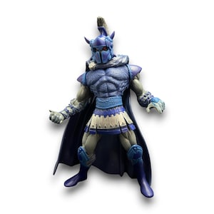 May include: A blue action figure of a warrior with a helmet, cape, and armour. The figure has a muscular physique and is posed with arms outstretched. The colour scheme includes shades of blue, white, and grey, with intricate detailing on the armour.